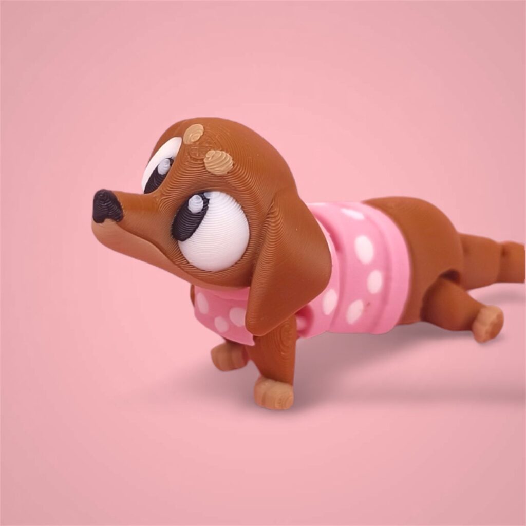 3D-gedruckte Printimon-Figur: kleiner brauner Dackel mit großen Kulleraugen und rosa Punkteshirt auf rosa Hintergrund.
